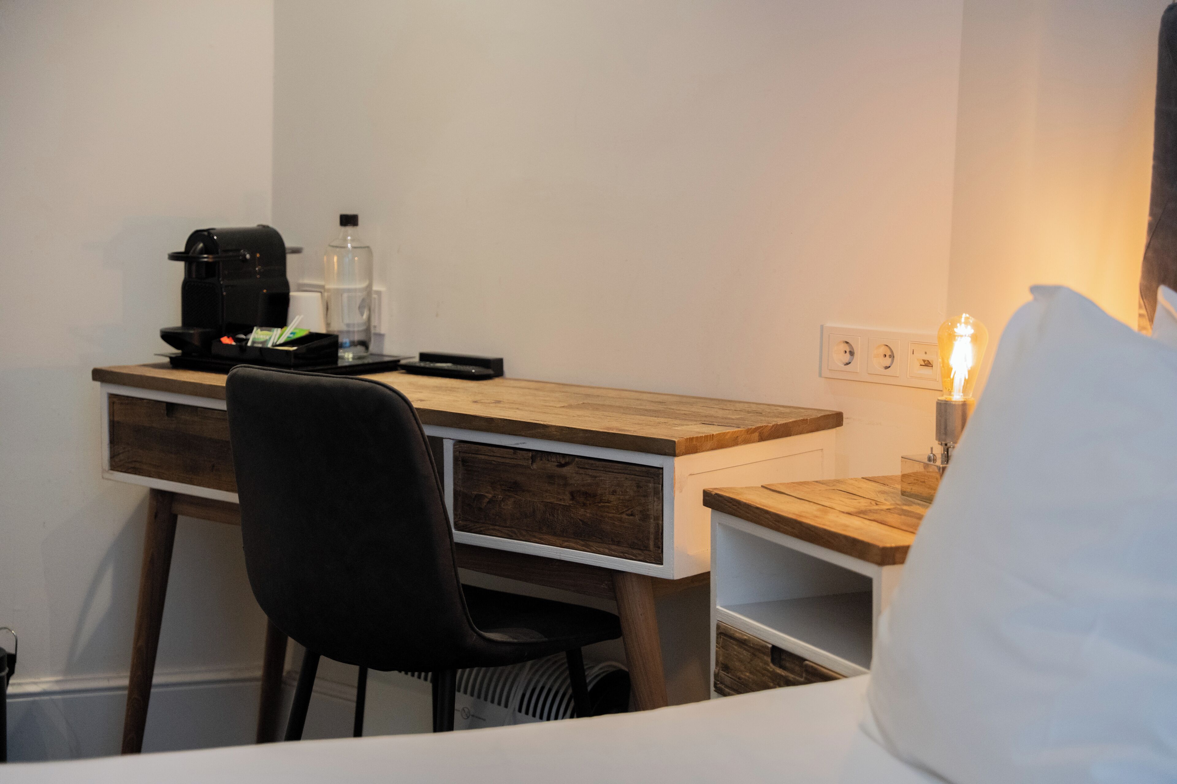 A hotel room with a wooden desk, a black chair, a sewing machine, and a bedside lamp.