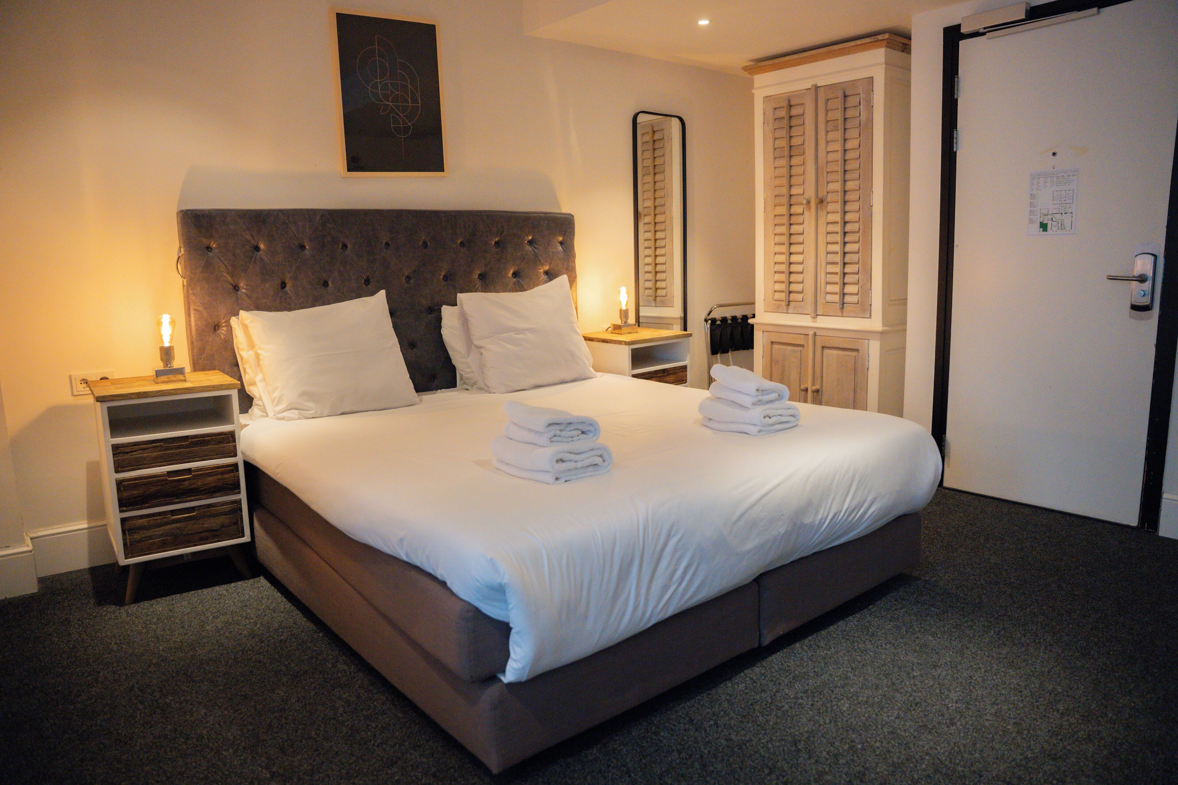 A neatly made bed with white linens and towels, a bedside table with a lamp, and a wardrobe with a mirror.