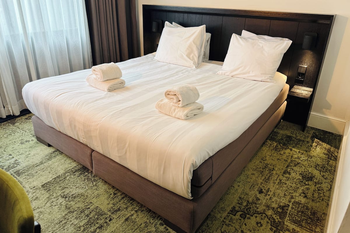 Une chambre d’hôtel avec un grand lit, deux oreillers, deux serviettes enroulées, une table de chevet avec une lampe et un tableau encadré au mur.