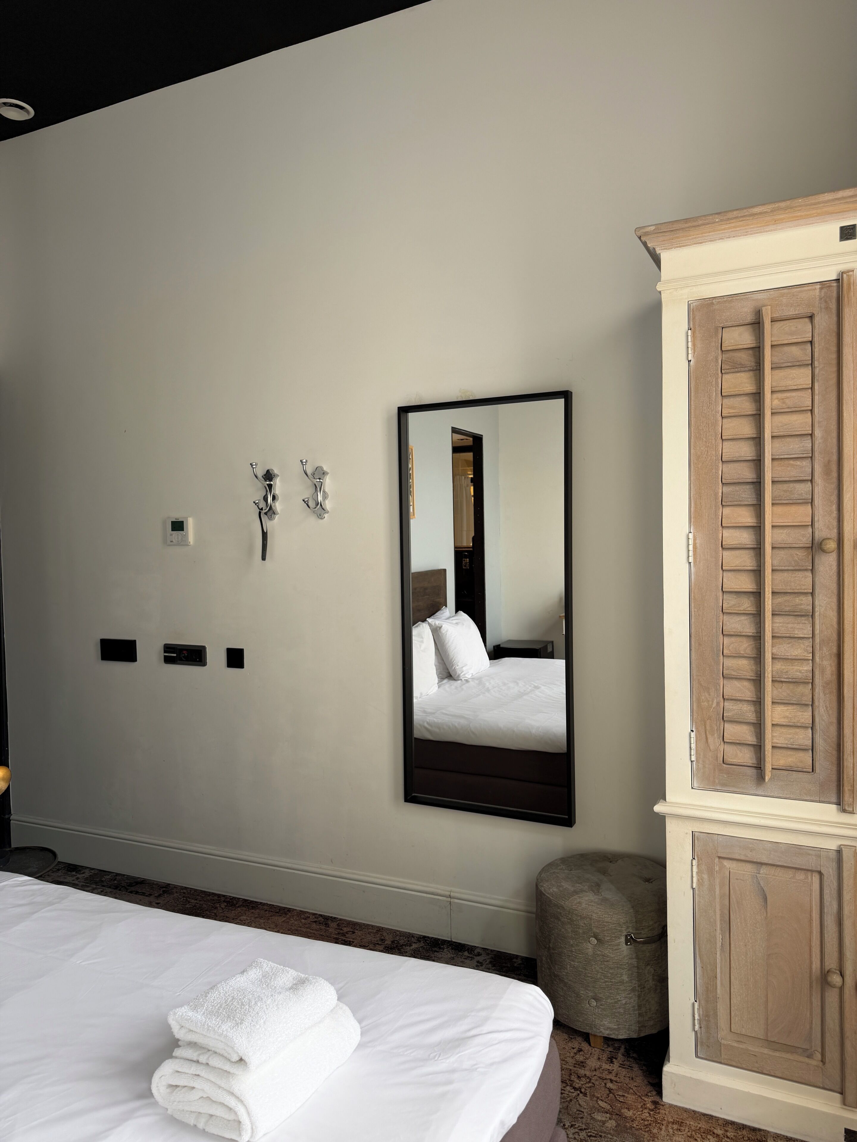 A hotel room with a bed, a mirror, a wooden wardrobe, and a small round ottoman.