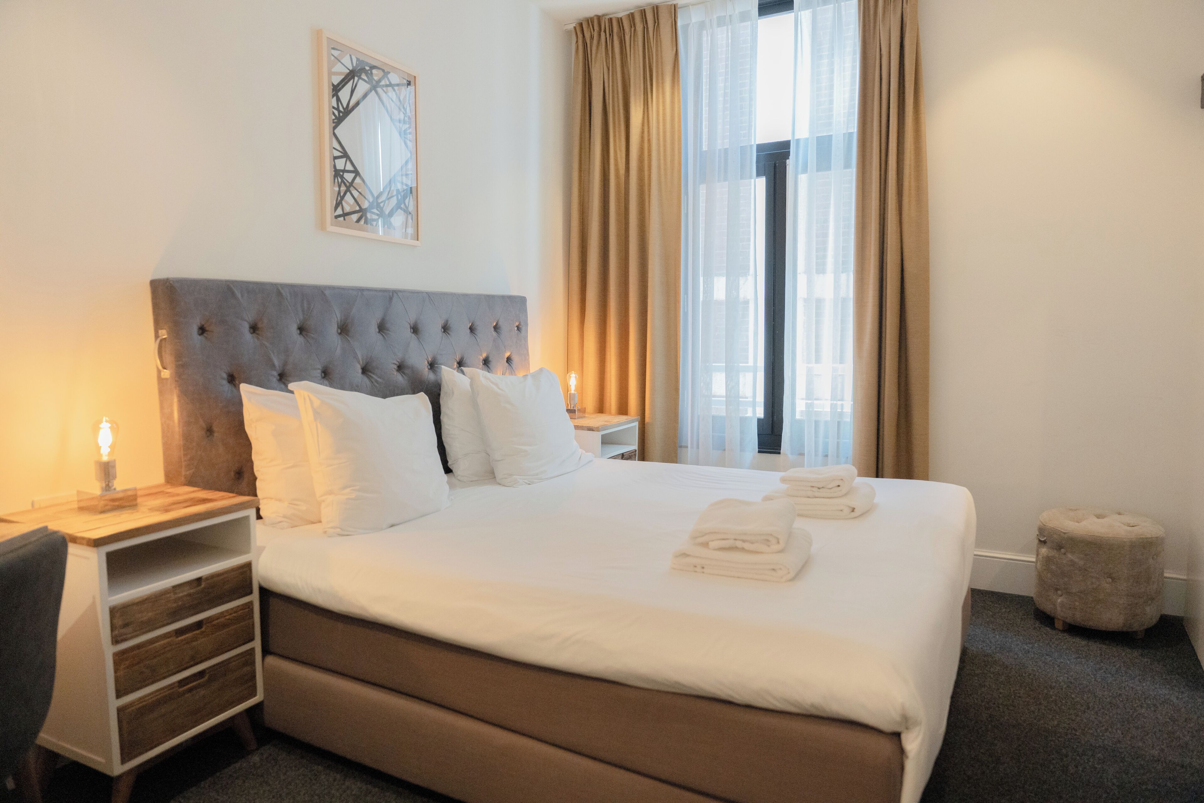 A neatly made bed with white linens, a tufted headboard, and a bedside table with a lamp.
