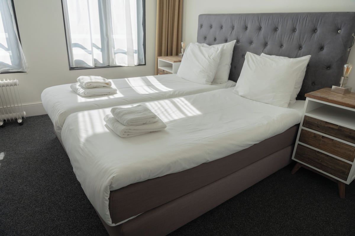 Un lit double avec du linge de lit blanc, une tête de lit rembourrée grise et une table de chevet avec une lampe.