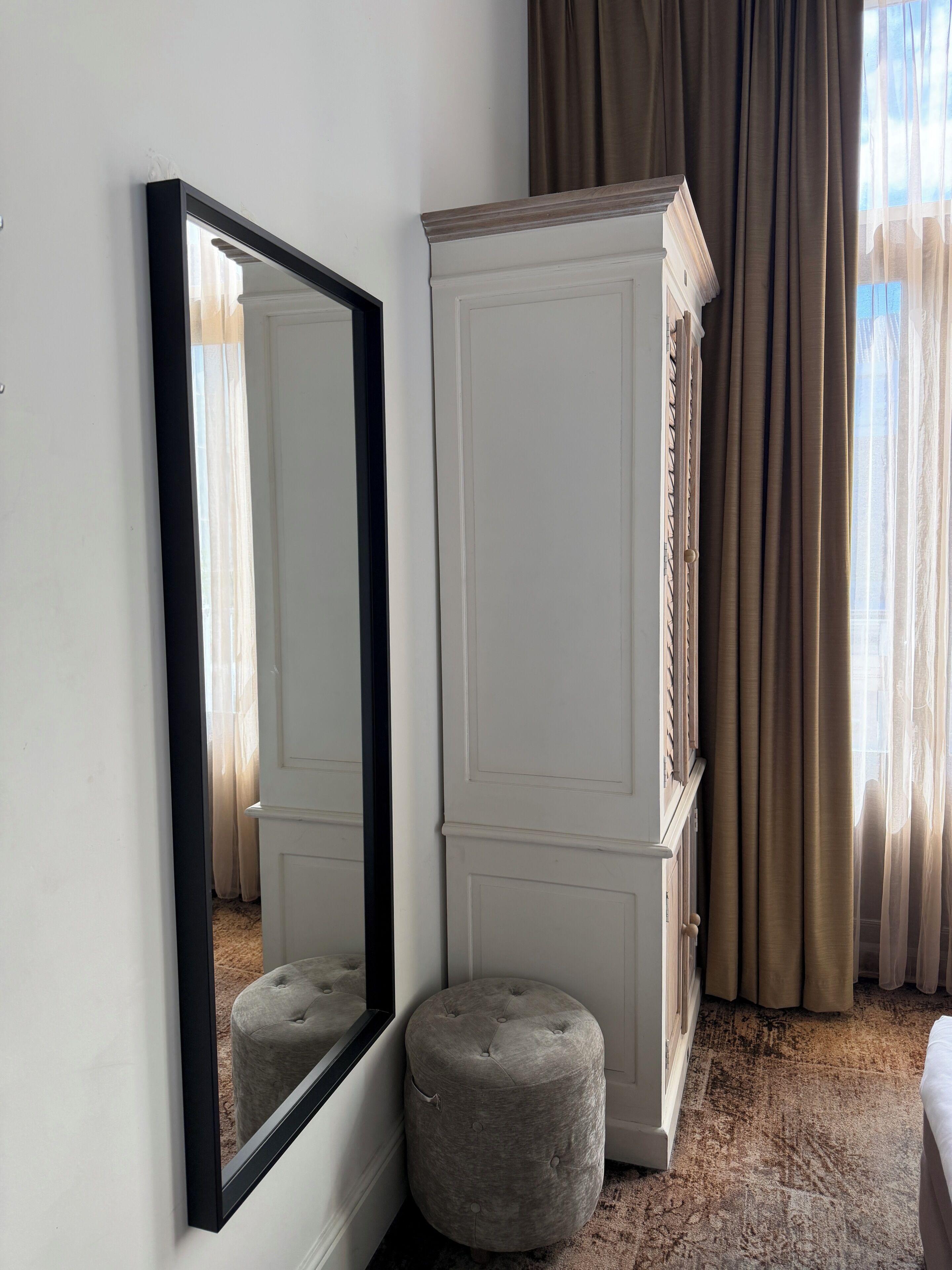 A room with a large mirror, a tall wardrobe, and a small ottoman.