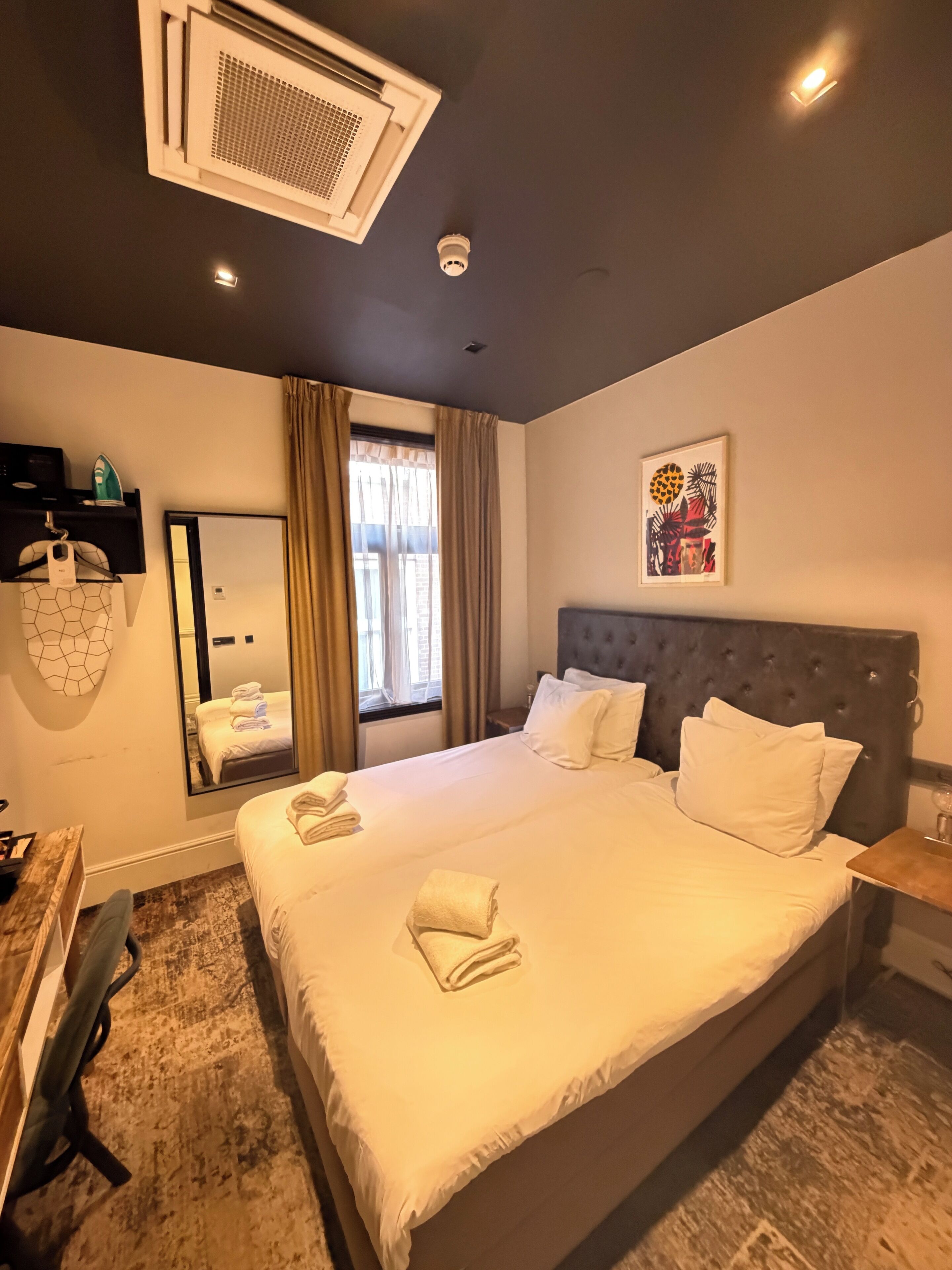 Hotelkamer met een groot bed, hoofdbord, nachtkastje, spiegel, raam met gordijnen en een kunstwerk aan de muur.