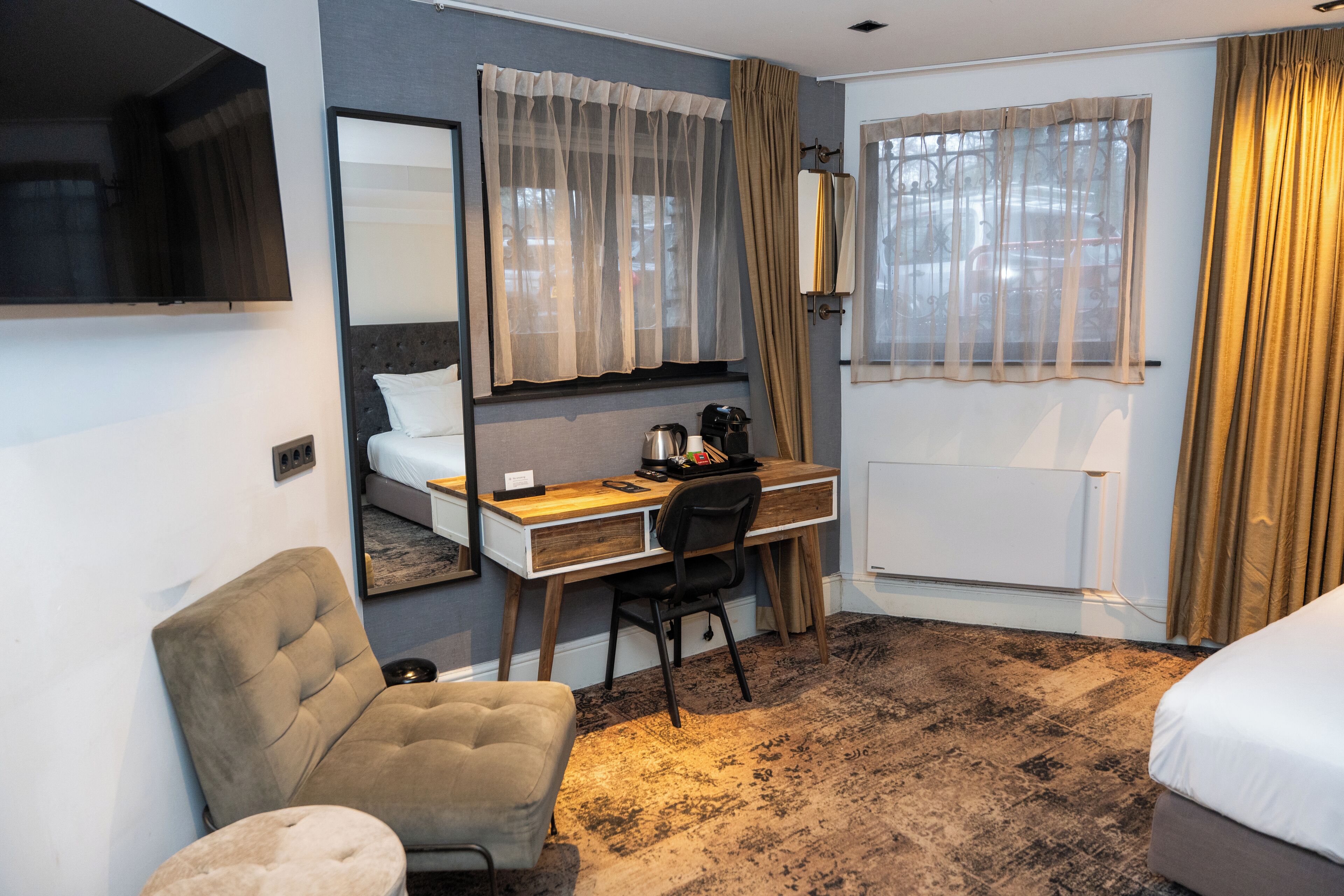 Quarto de hotel com cama, escrivaninha, poltrona, sofá e uma grande janela com cortinas.