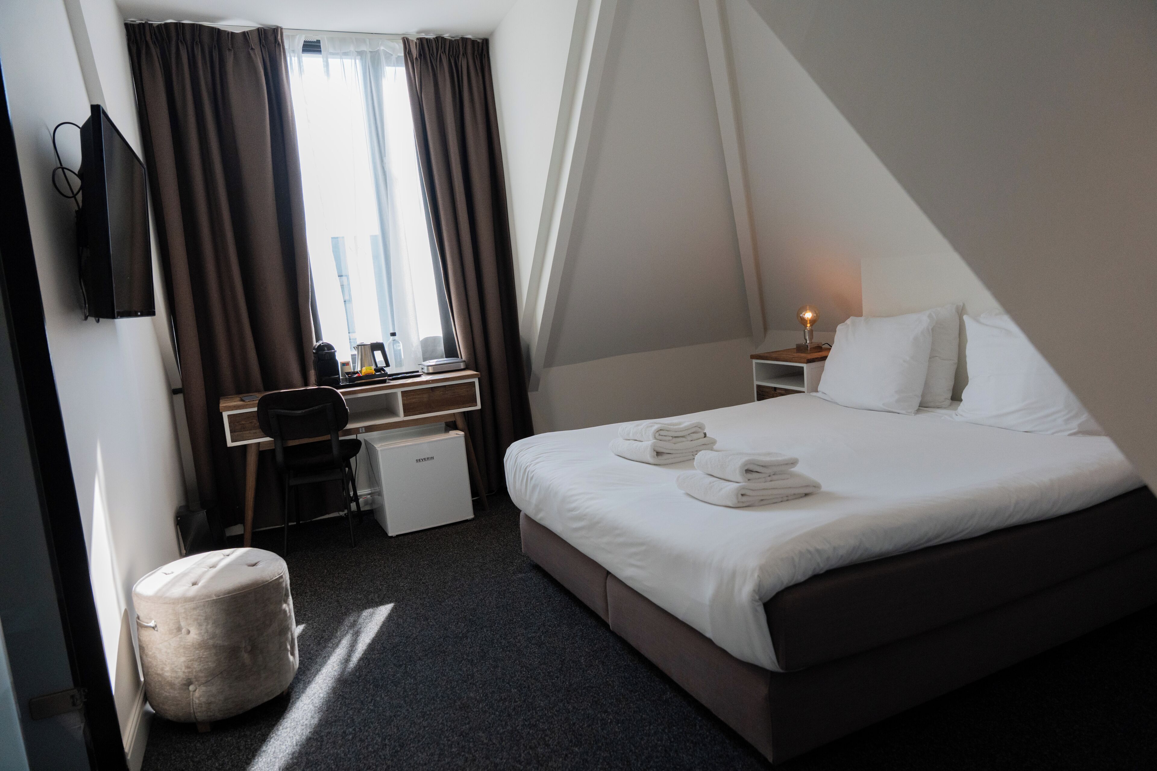 A hotel room with a bed, a desk, a chair, a small ottoman, a TV, and a window with curtains.