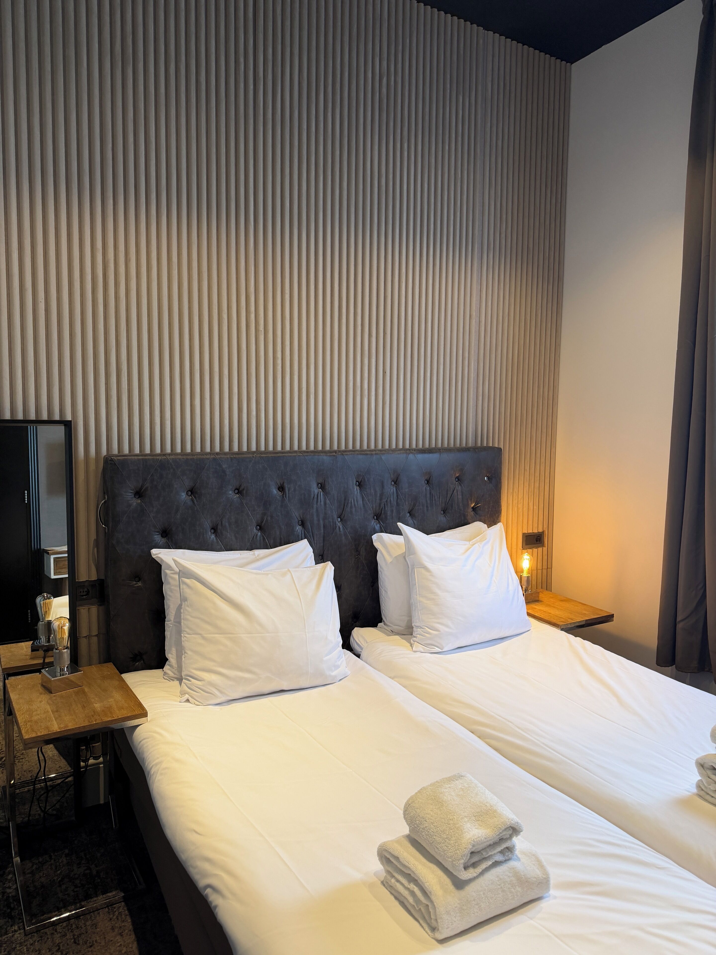 A hotel room with two beds, a dark upholstered headboard, white bedding, a bedside table with a lamp, and a wall with vertical blinds.