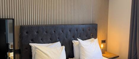 A hotel room with two beds, a dark upholstered headboard, white bedding, a bedside table with a lamp, and a wall with vertical blinds.