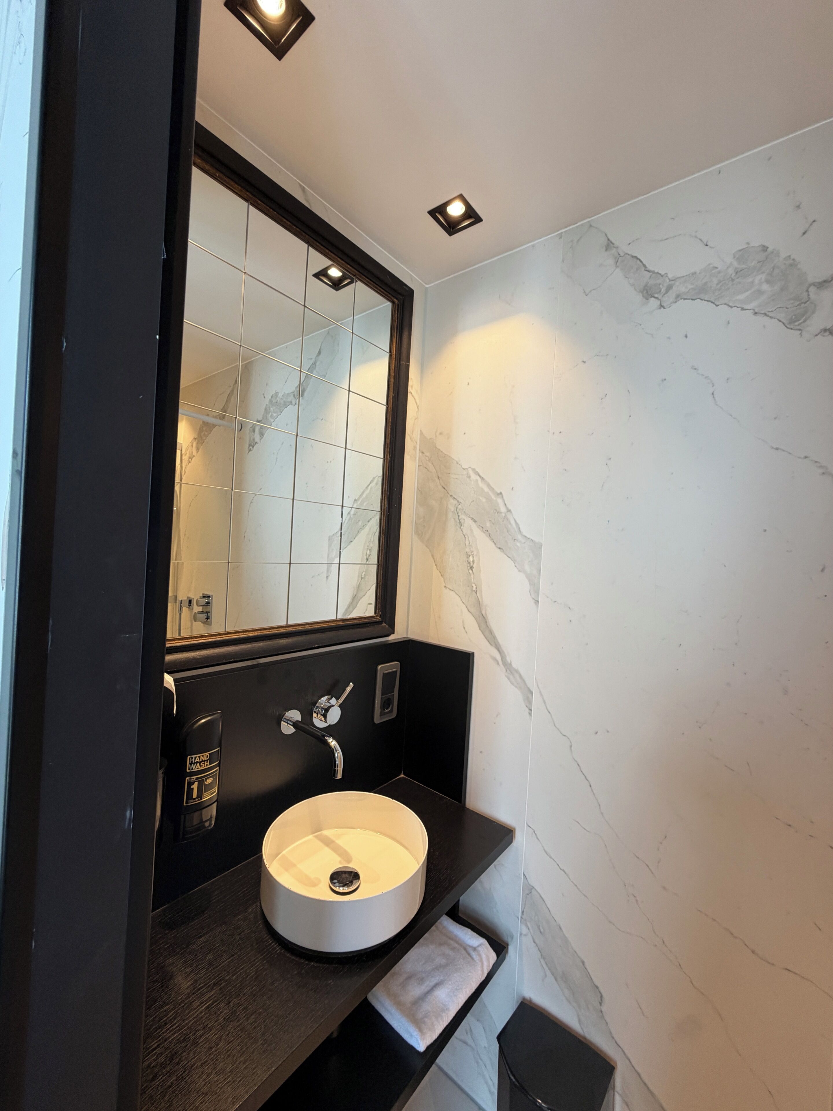 Un baño moderno con pared de mármol, un lavabo redondo, estación de lavado de manos y un espejo sobre el lavabo.
