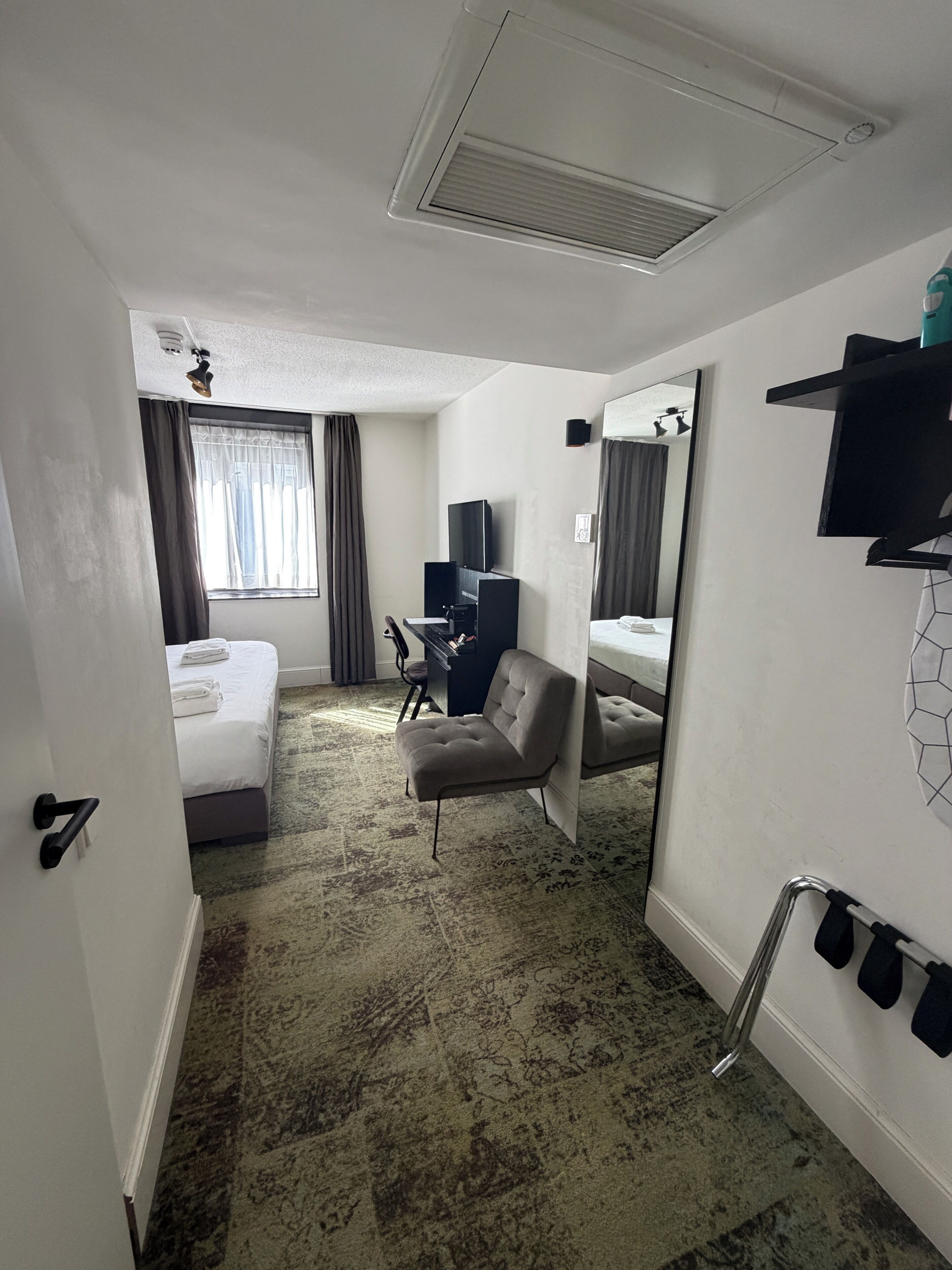A hotel room with a bed, a chair, a desk, a television, a mirror, and a shower area.