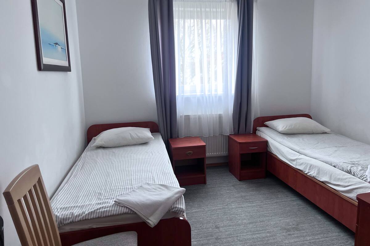A small, clean room with two single beds, a chair, a small table, and a window with curtains.