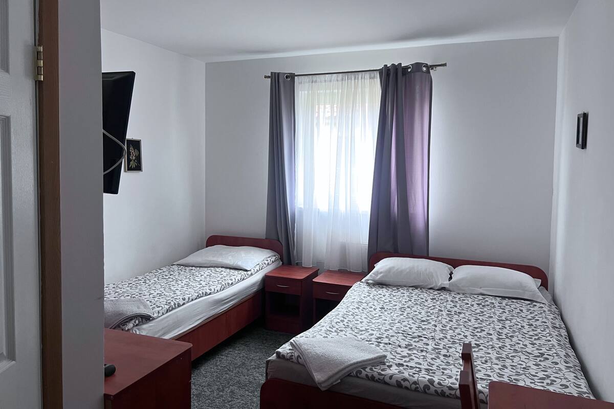 A small, clean room with two single beds, a bedside table, a nightstand, and a window with curtains.
