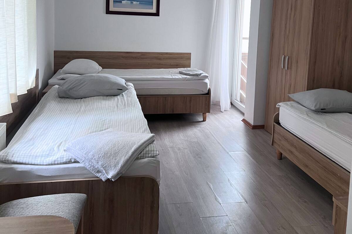 A room with two single beds, a wooden wardrobe, a framed picture on the wall, and a window with curtains.