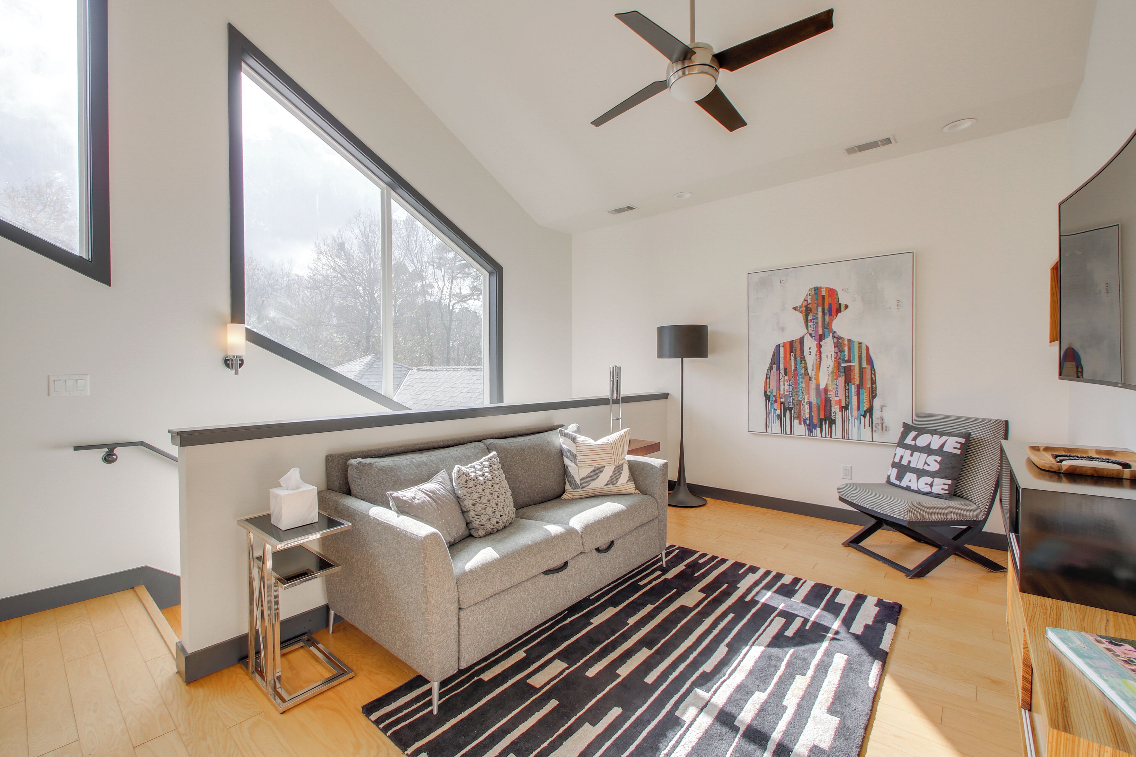 A modern living room with a grey sofa, a black and white rug, a ceiling fan, and a large window.