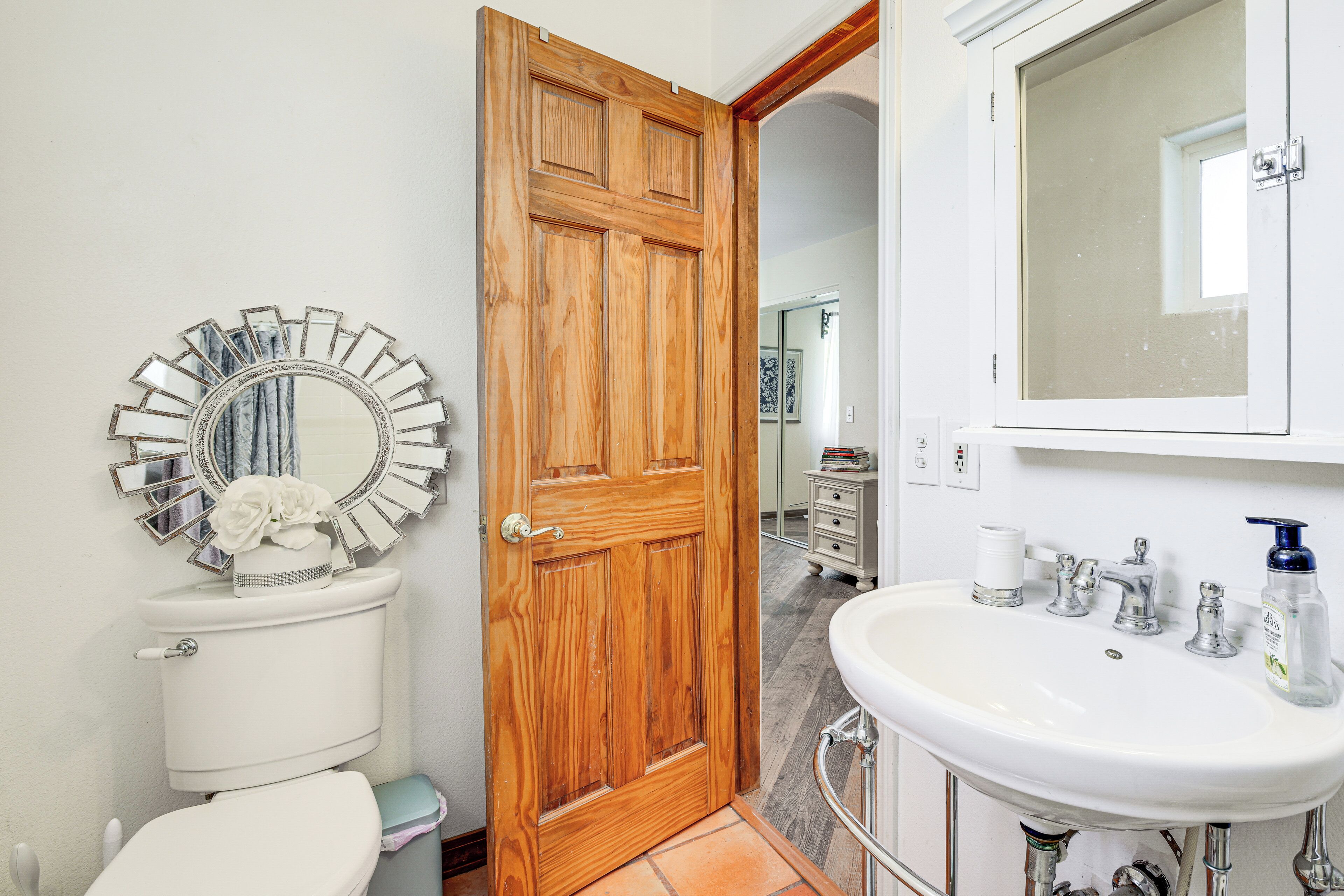 A bathroom with a wooden door, a white toilet, a round mirror with a decorative frame, and a white sink with a mirror above it.