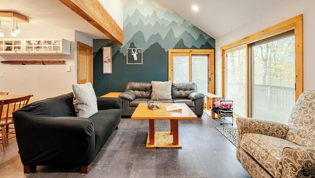 A cozy living room with a wooden coffee table, two sofas, a dining area, and a large window with blinds. - Ski-in/ski-out Camelback Mtn Retreat w/ Hot Tub (Tannersville)