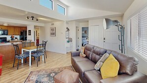 A spacious living area with a brown leather sofa, a dining table with chairs, a kitchen in the background, and a large window with blinds. - 1 BR Condo in Tucson, Mountain Views (Tucson)