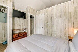 Un dormitorio con una cama, un televisor montado en la pared, un aparador de madera y una puerta que da a otra habitación.