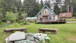 A cabin with a green roof, a deck, and a large log pile in a forested area. - Pacific View Home: Hot Tub & Ocean/valley Views! (Langlois)