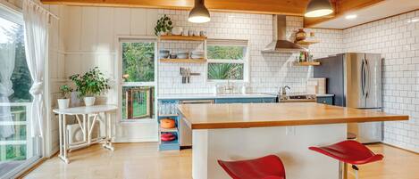 A modern kitchen with a central island, wooden beams, white tiled walls, and a large window.