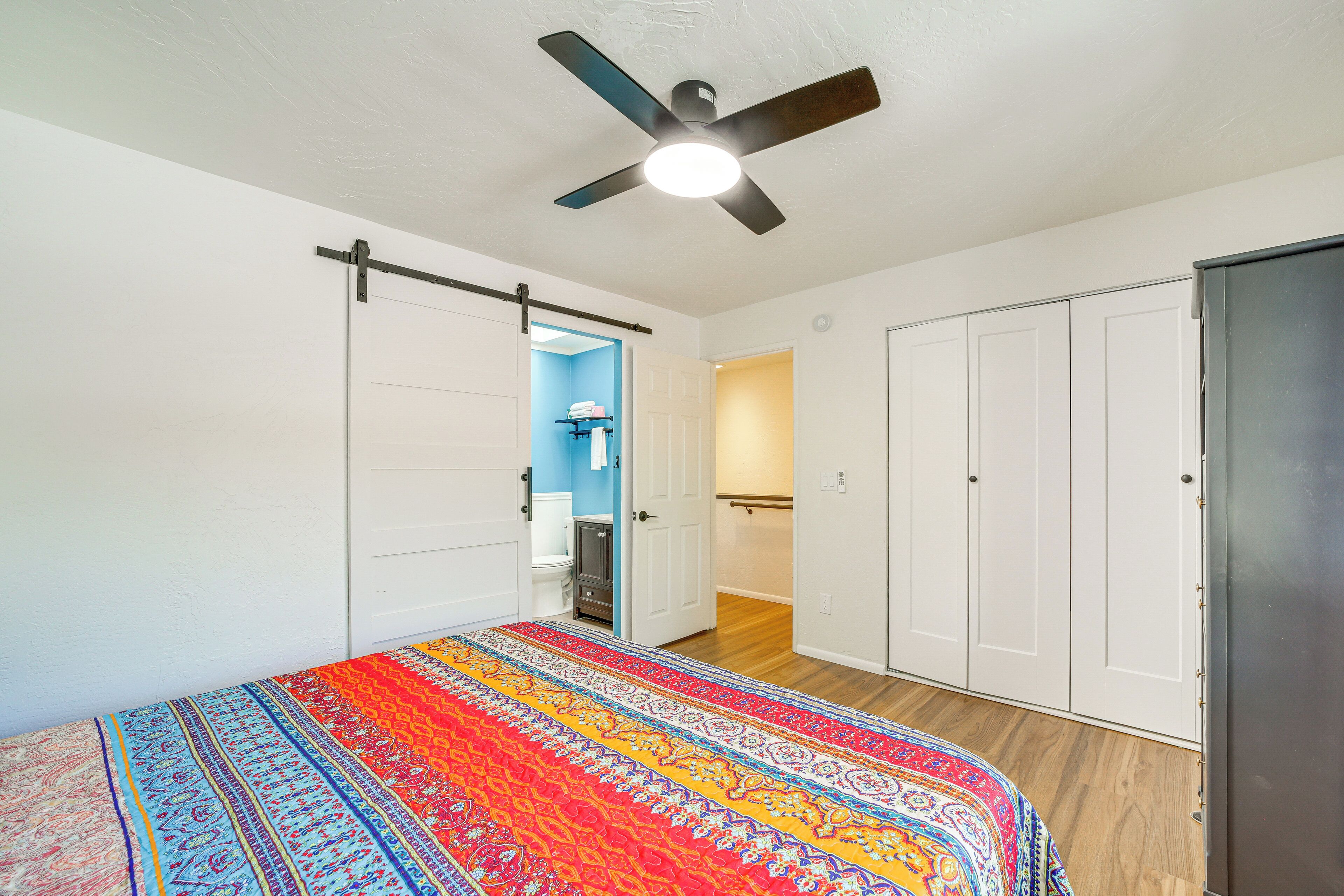 Une chambre à coucher comprenant un lit, un ventilateur de plafond, des armoires blanches et une porte donnant sur une salle de bain.