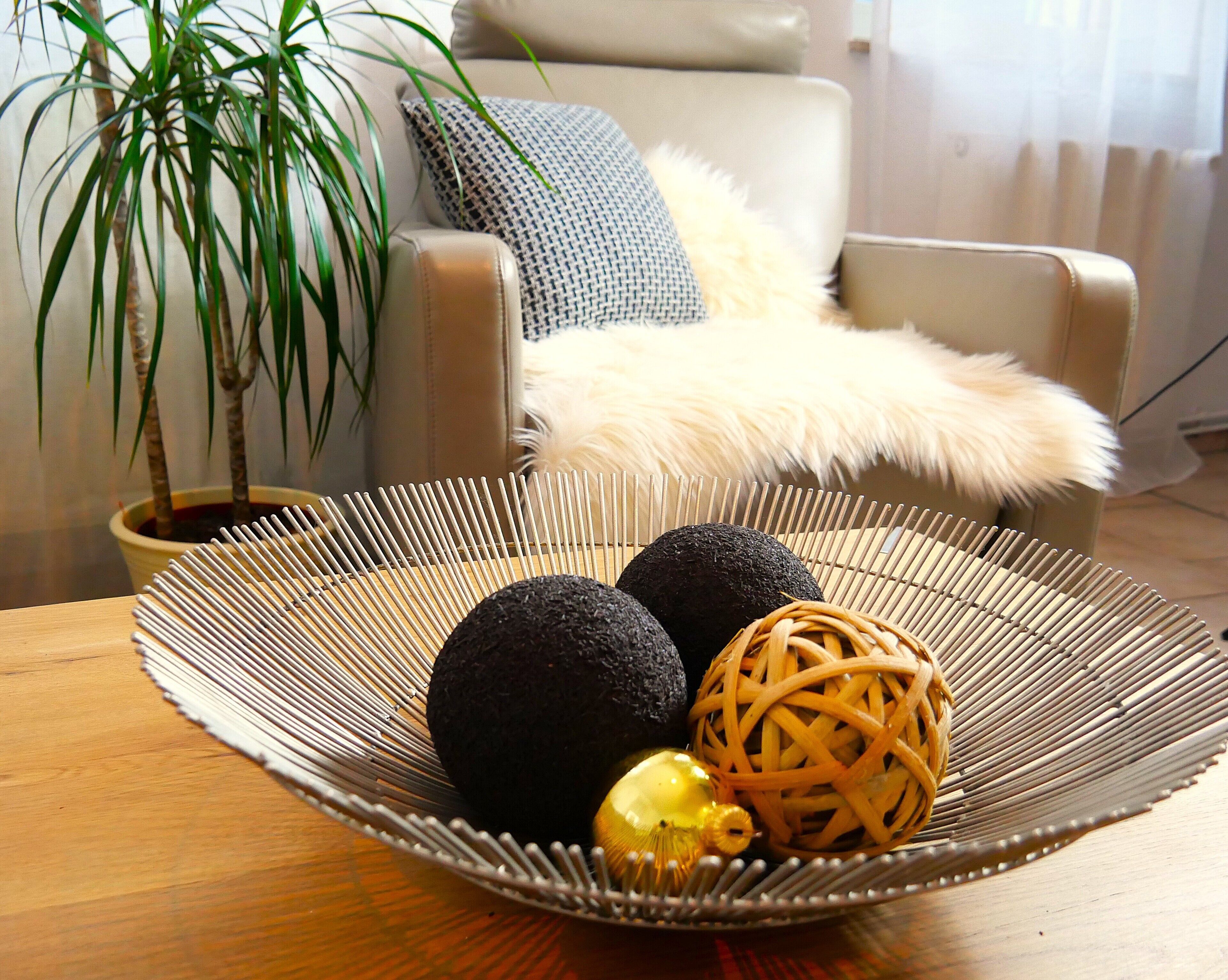 A round wicker tray with decorative items on a wooden table, a potted plant, and a sofa with a fluffy throw blanket in a living room.