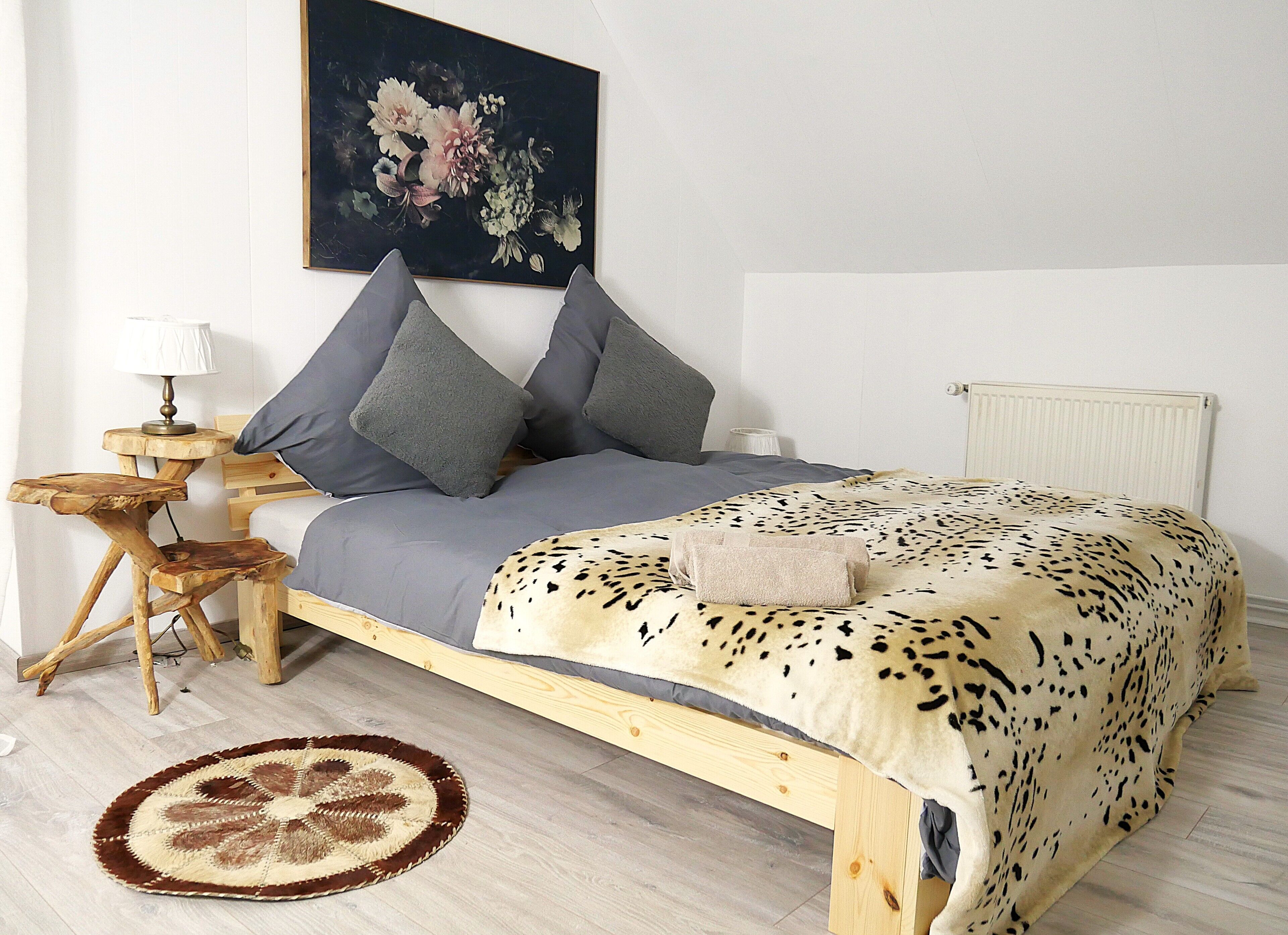 A bedroom with a wooden bed, grey bedding, a leopard print blanket, a round rug, a wooden nightstand with a lamp, and a floral painting on the wall.