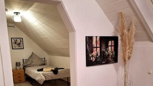 Um quarto de sótão acolhedor com teto inclinado, uma cama com travesseiro, uma mesinha de cabeceira, um abajur e um quadro com flores.