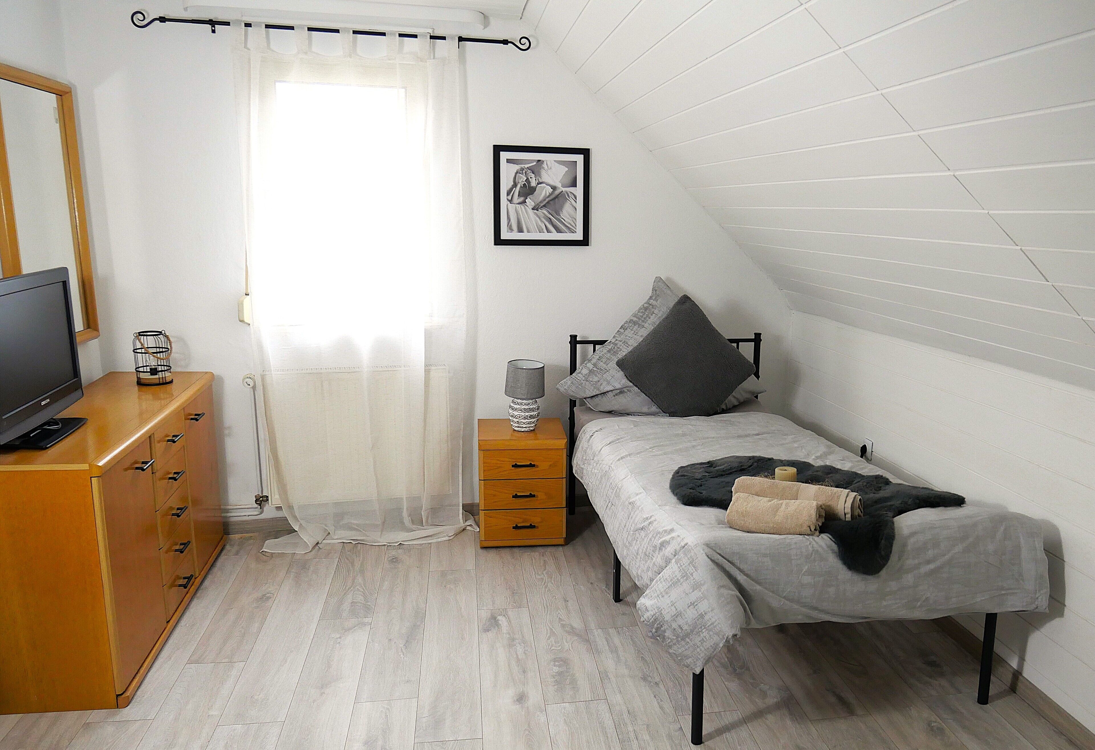 Une chambre à coucher comprenant un lit, une commode, un téléviseur, une fenêtre avec des rideaux et un tableau encadré accroché au mur.