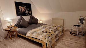Quarto com teto inclinado, cama com manta estampada, mesa de cabeceira com abajur, uma cadeira e um radiador.