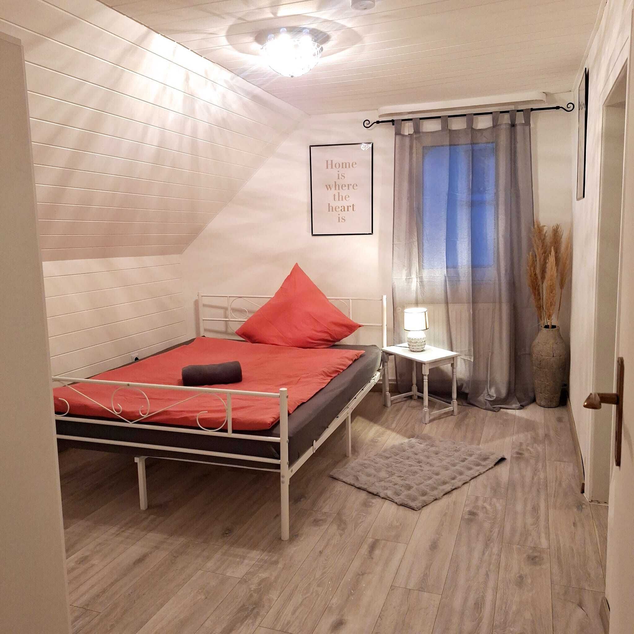 A small, cozy bedroom with a sloped ceiling, a single bed with red bedding, a bedside table with a lamp, a window with sheer curtains, and a decorative sign on the wall.