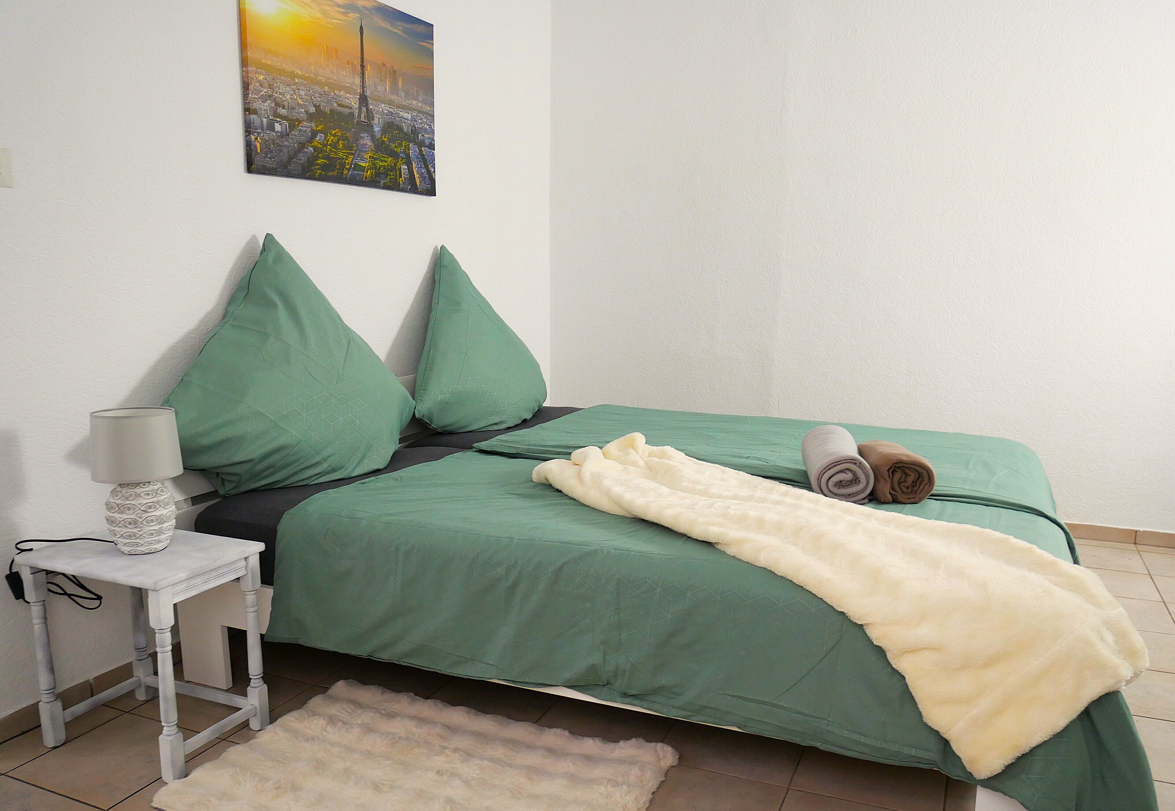 A neatly made bed with green bedding and a white throw blanket, a small bedside table with a lamp, and a fluffy rug on the floor.