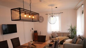 Uma sala de estar moderna com uma mesa de jantar de madeira, cadeiras escuras, um sofá cinza e uma grande planta em vaso.