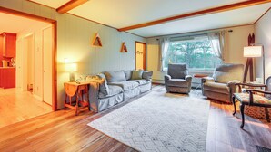 A spacious living room with a wooden floor, a grey sofa, a brown armchair, and a small table with a lamp.