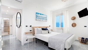 Ein modernes Schlafzimmer mit Bett, kleinem Tisch, Spiegel und Meerblick durch ein Fenster.