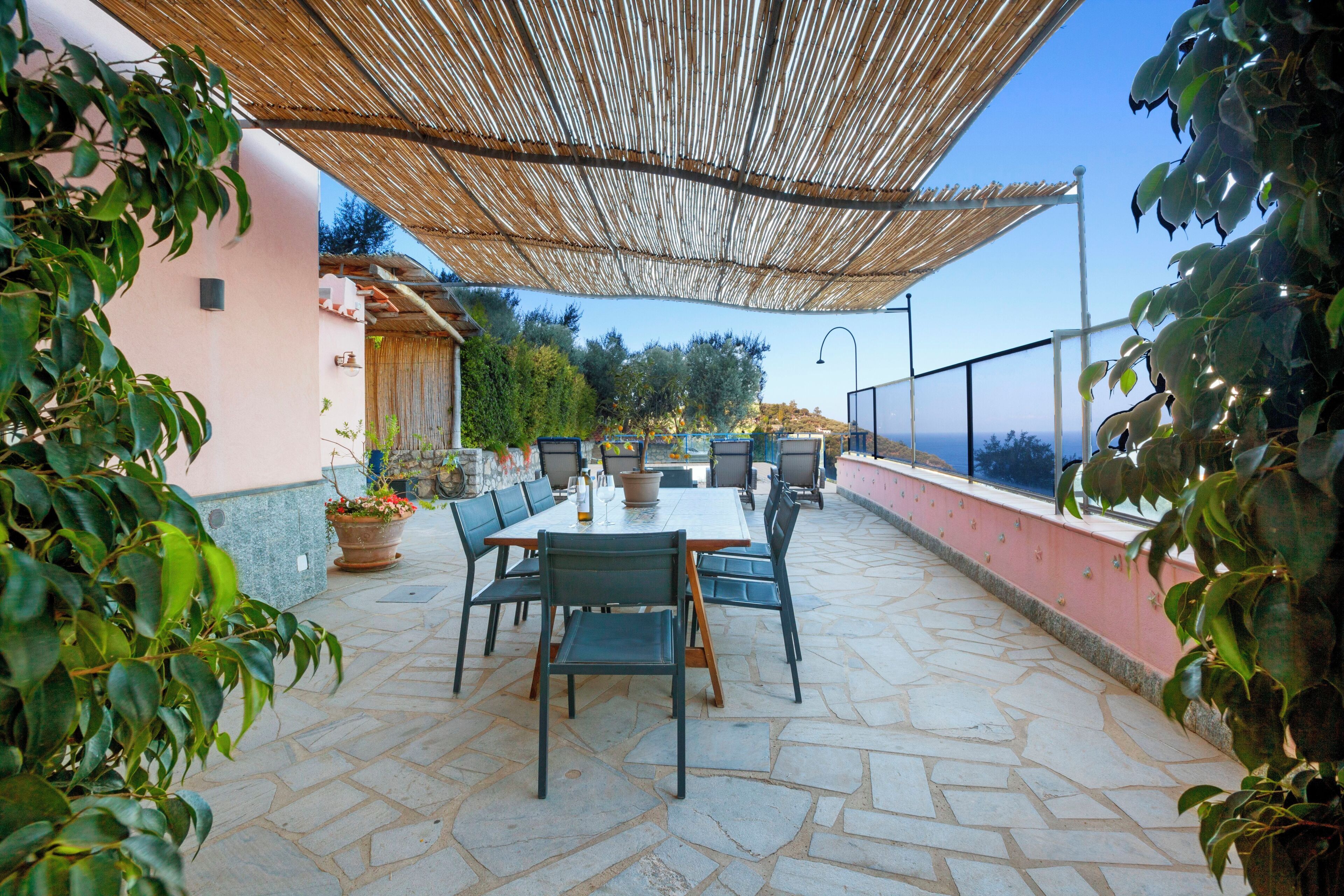 Une terrasse extérieure avec un sol en pierre, une table entourée de chaises et une pergola offrant de l’ombre.