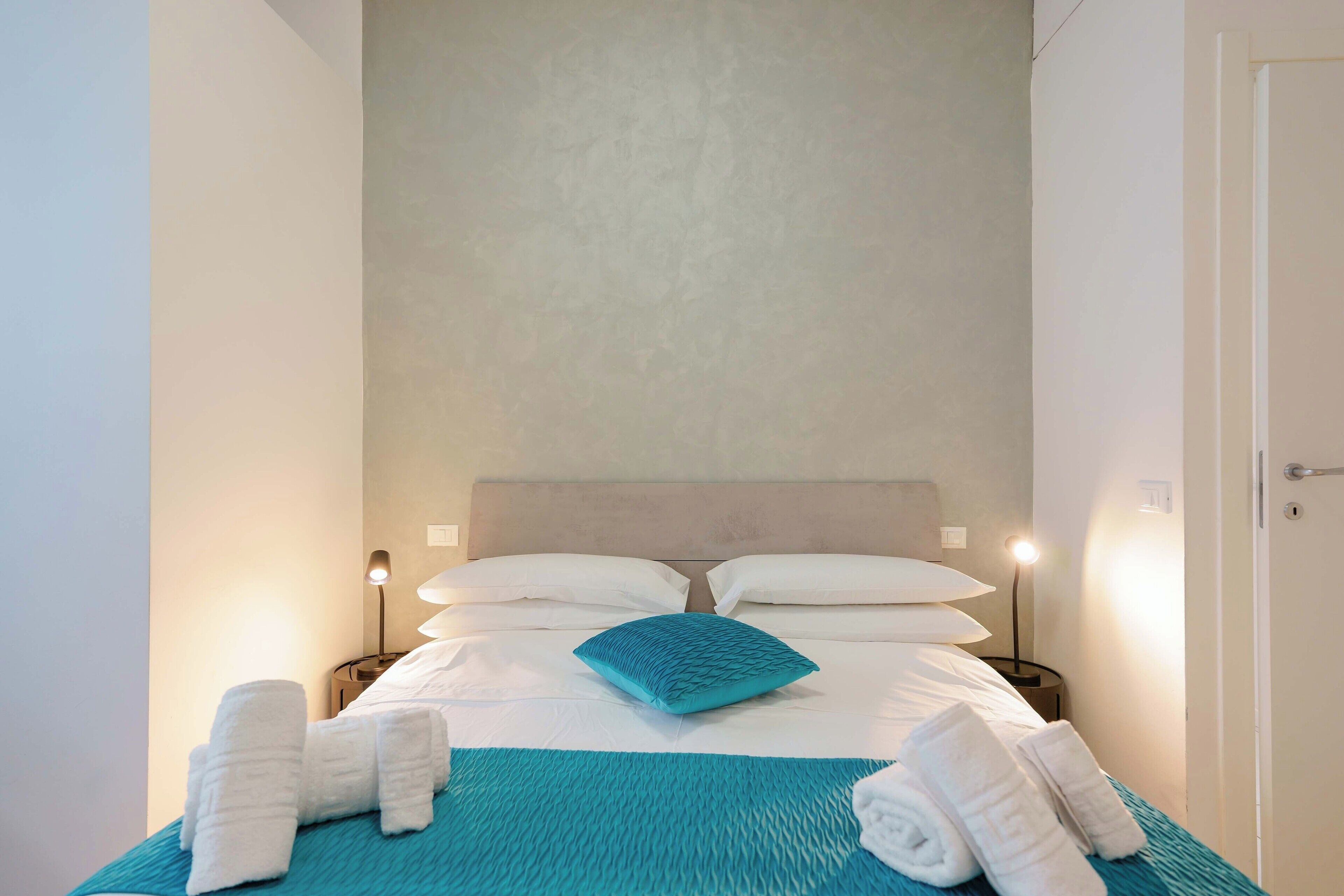 Una cama bien tendida con sábanas blancas y un cojín turquesa, dos lámparas de noche y toallas enrolladas sobre la cama.