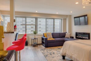 A modern hotel room with a bed, a sofa, a red chair, a small table with a plant, and a TV mounted on the wall. - Cozy 12th-floor Studio w/ Lake Erie Views! (Lakewood)