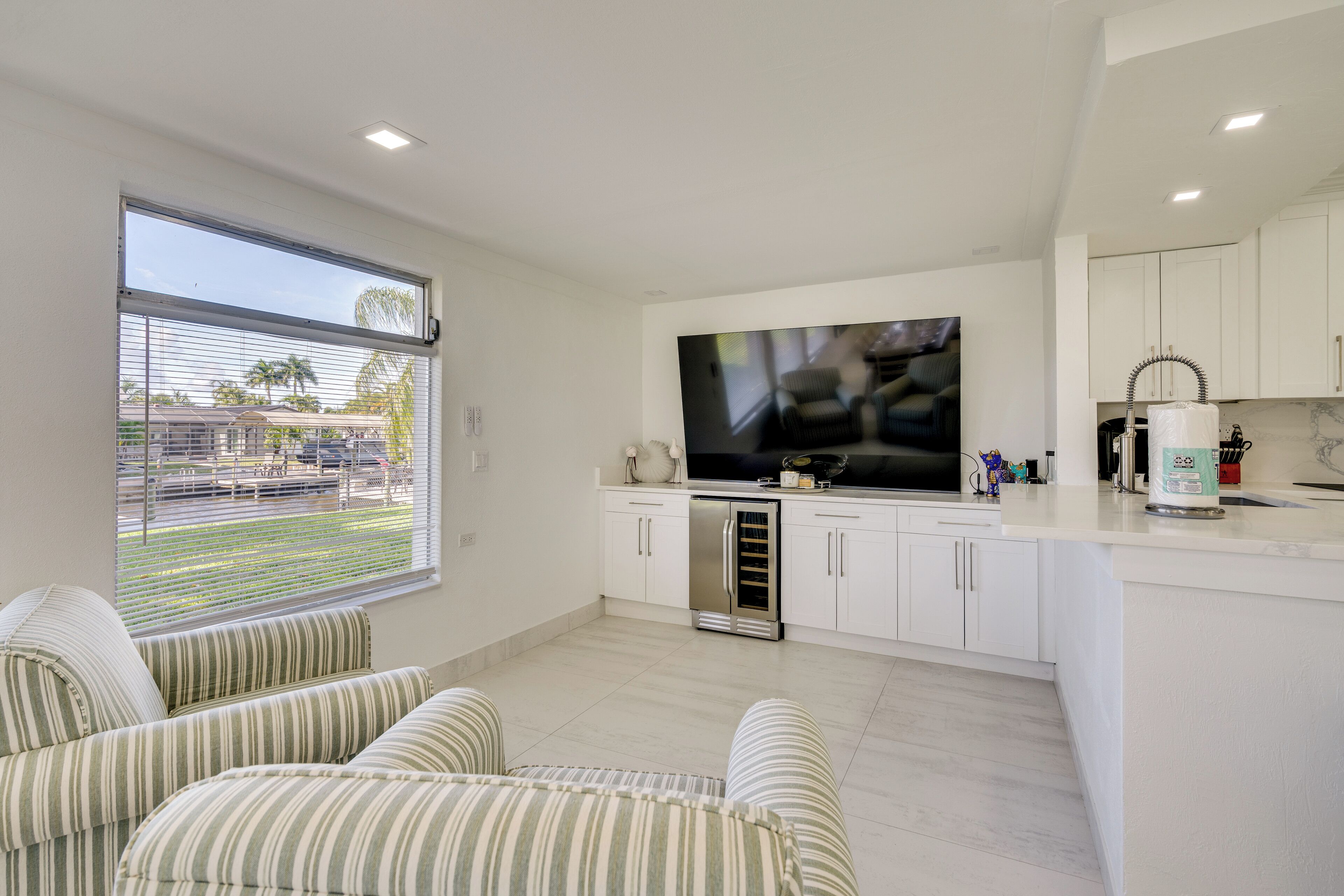 A modern kitchen with white cabinets, a large TV, and a view of a train and palm trees outside.