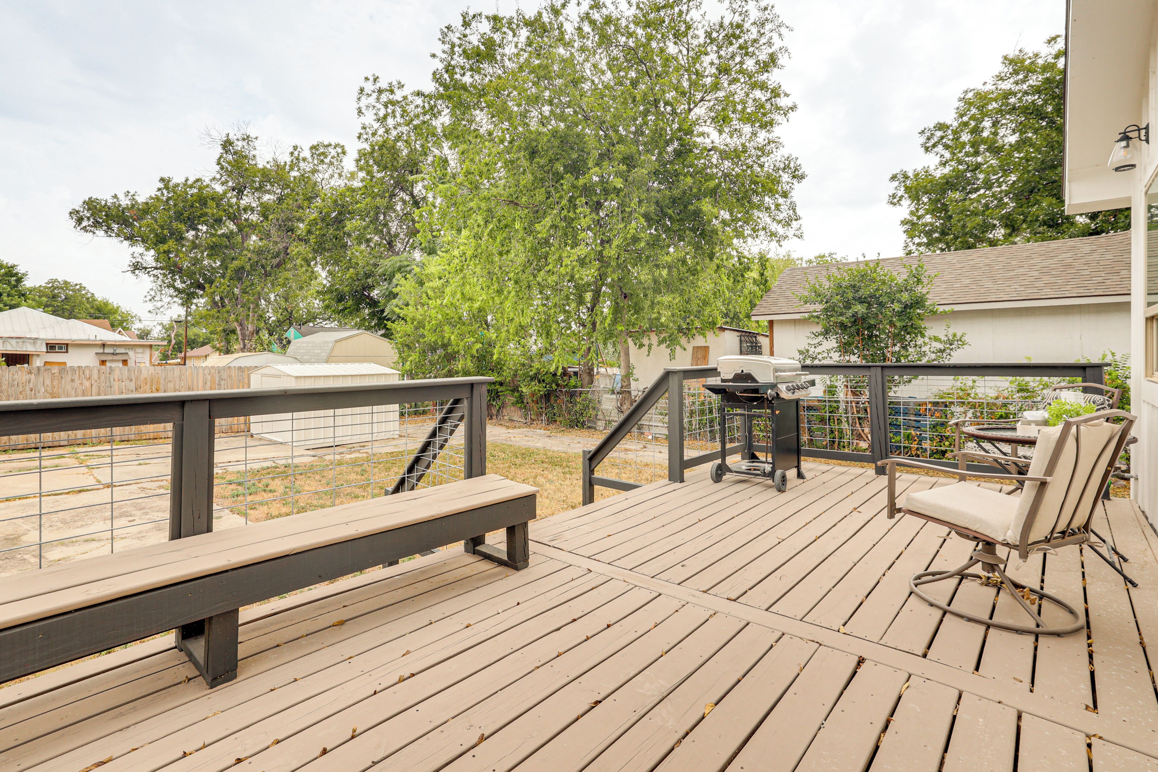 Una terraza de madera con un banco y una silla, una parrilla y un área cercada con árboles.