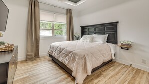 Une chambre à coucher avec un lit, un plancher en bois, un ventilateur de plafond, une fenêtre avec des rideaux et une table de chevet sur laquelle est posée une plante.