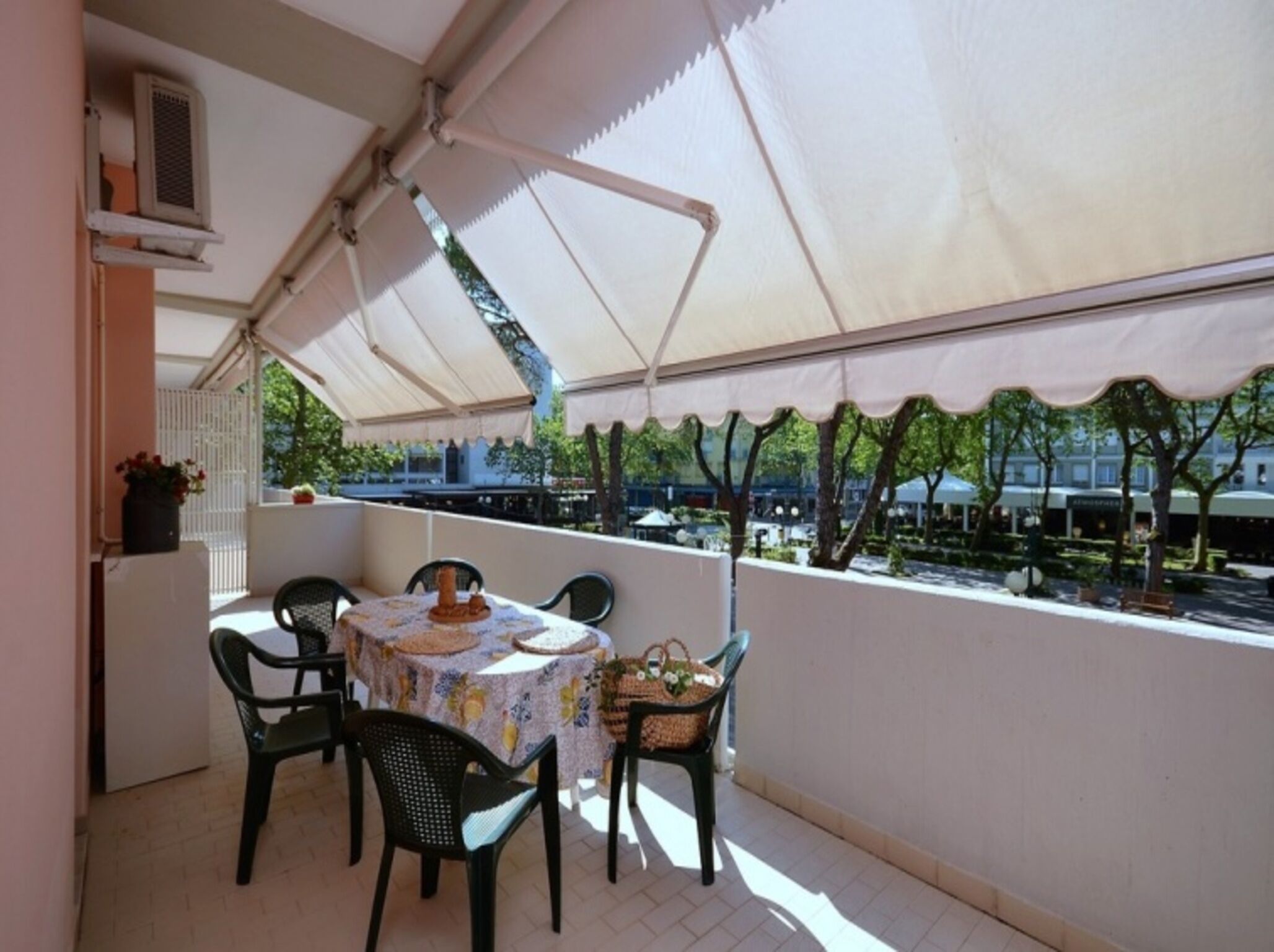 A balcony with a table and chairs, covered by a white canopy, overlooking a garden with trees and a building in the background.