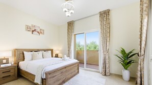 Ein Schlafzimmer mit einem hölzernen Bett, einem Nachttisch, einem Beistelltisch, einem großen Fenster mit Vorhängen und einer Topfpflanze.