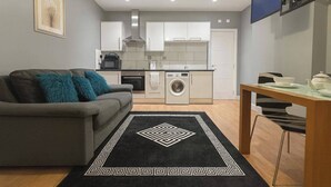 A compact living space with a grey sofa, a washing machine, a dining table, and a TV mounted on the wall. - Charming High Wycombe Apartment (High Wycombe)