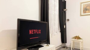 A room with a TV displaying the Netflix logo, a small white table with a plant, a floor lamp, and a framed picture on the wall. - Charming High Wycombe Apartment (High Wycombe)