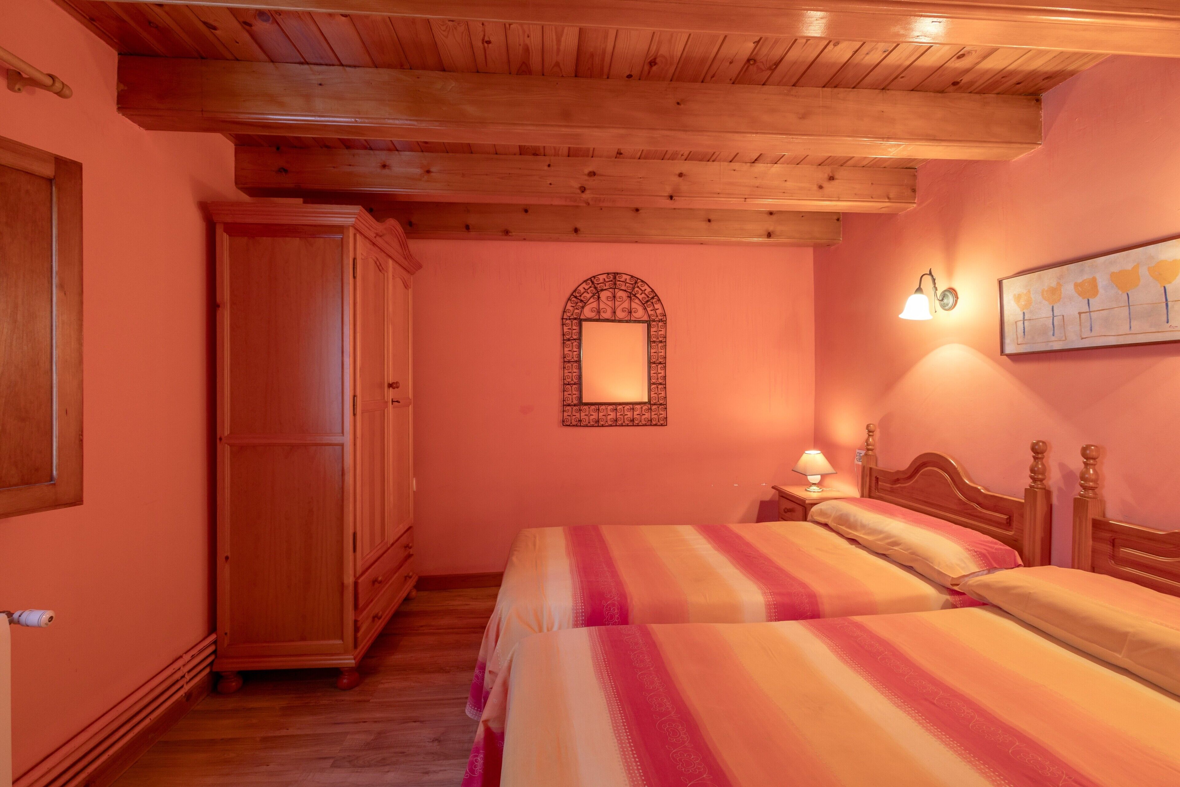 Quarto com duas camas, guarda-roupa de madeira, espelho com moldura decorativa e uma luminária fixada na parede.