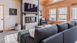 Ein gemütliches Blockhäuschen-Interieur mit einem großen grauen Sofa, einem Kamin, einem Fernseher und einem Fenster mit der Aufschrift 'SUND'.