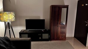 Una habitación con una televisión sobre un soporte negro, un armario de madera con espejo, una lámpara con pantalla estampada y una alfombra con motivos en el suelo.