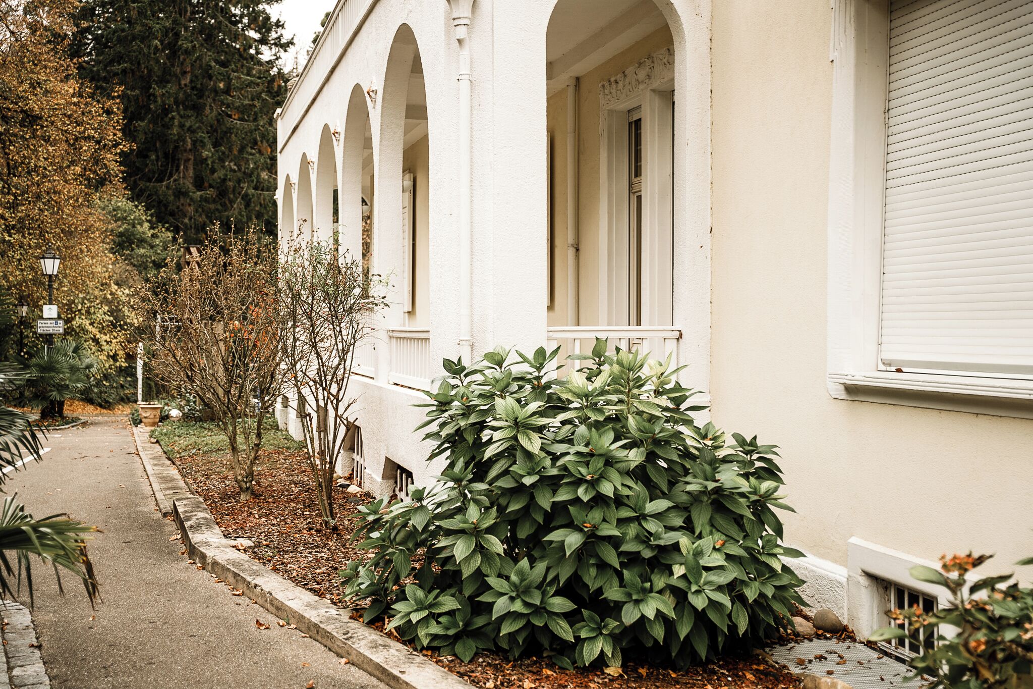 A building with a white facade, arched windows, and a small garden area with green shrubs.