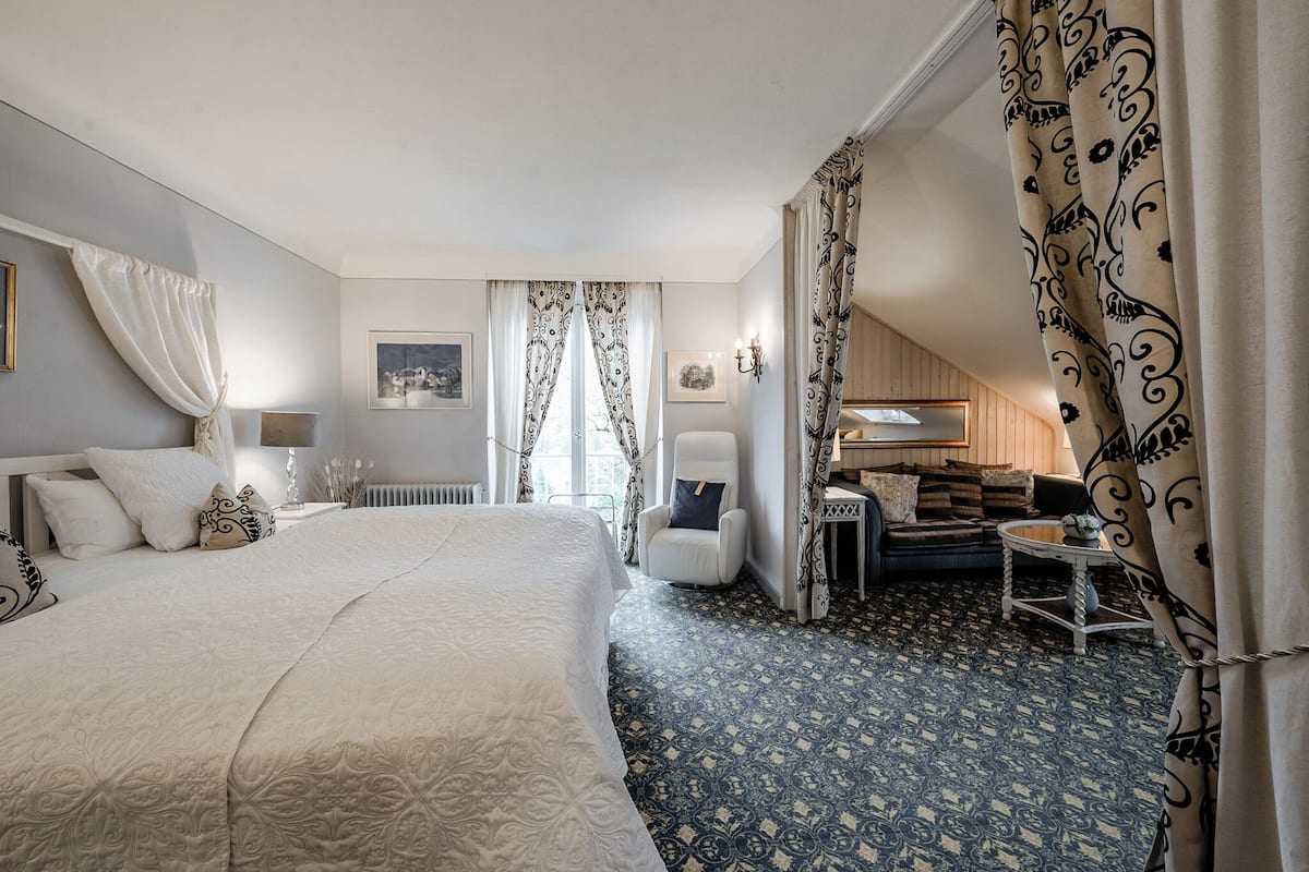 A hotel room with a large bed, a small sofa, a side table, and a chair. There are curtains, a picture on the wall, and a lamp.